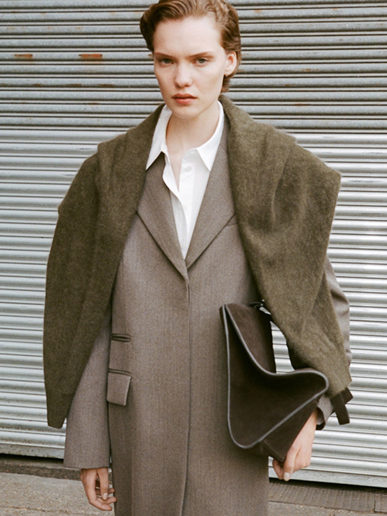 Person in a long gray coat with a dark green shawl over the shoulders, holding a dark handbag, standing in front of a corrugated metal wall.