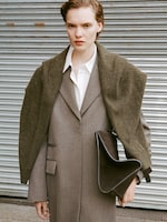 Placeholder for: Person in a long gray coat with a dark green shawl over the shoulders, holding a dark handbag, standing in front of a corrugated metal wall.