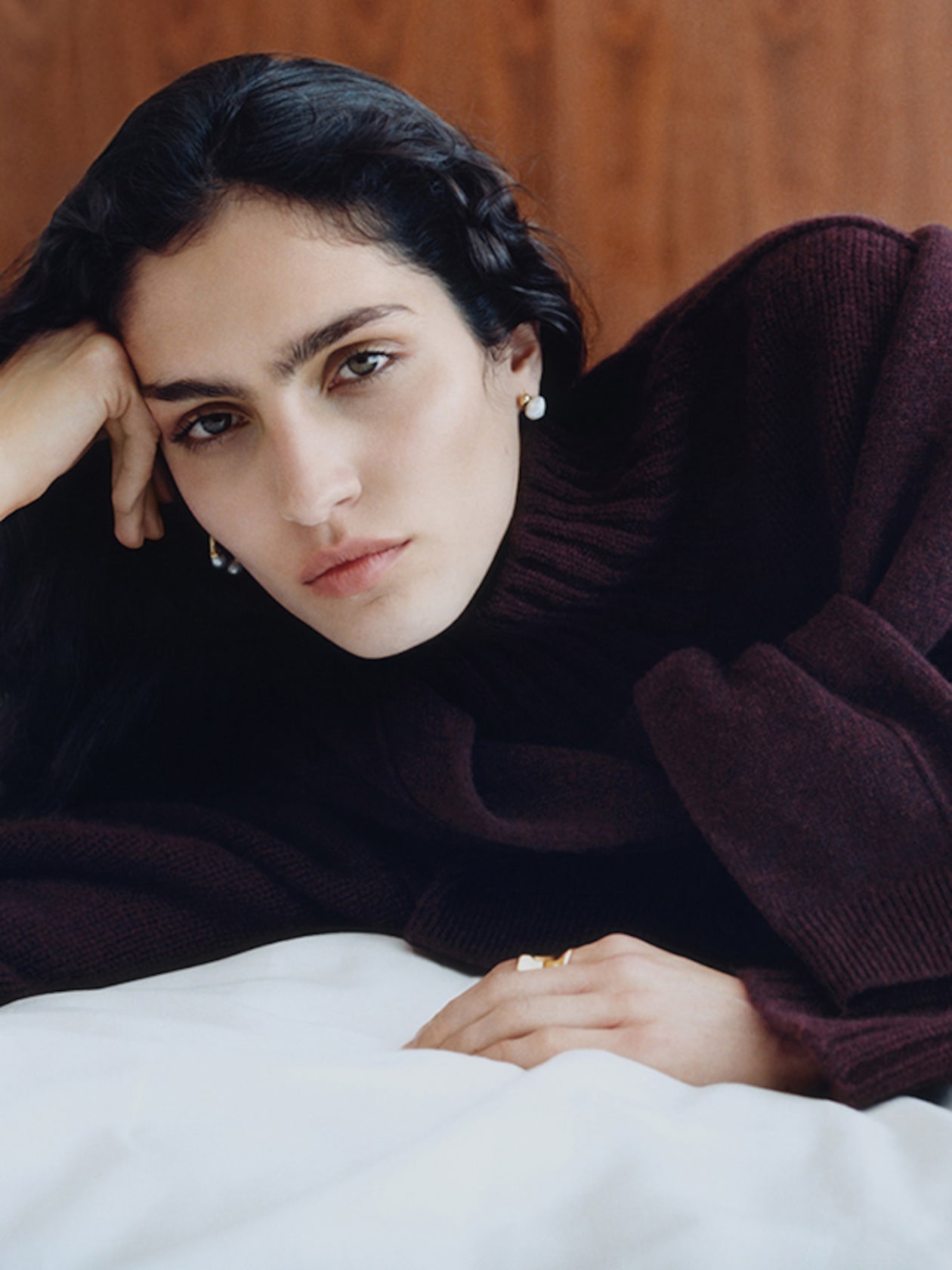 Person with long dark hair and pearl earrings, wearing a burgundy sweater, rests head on hand against a wooden backdrop.
