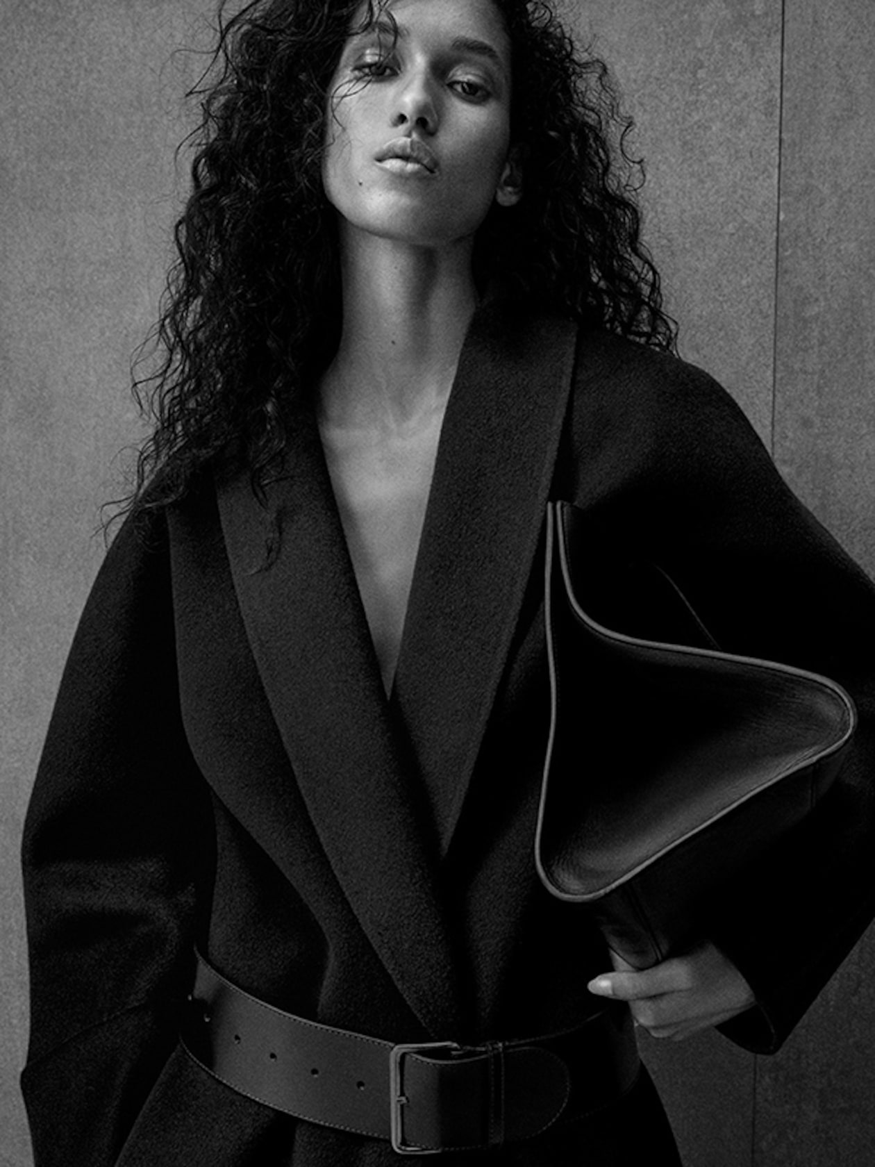Black and white photo of a person with curly hair, wearing a belted coat, holding a triangular clutch, against a plain background.
