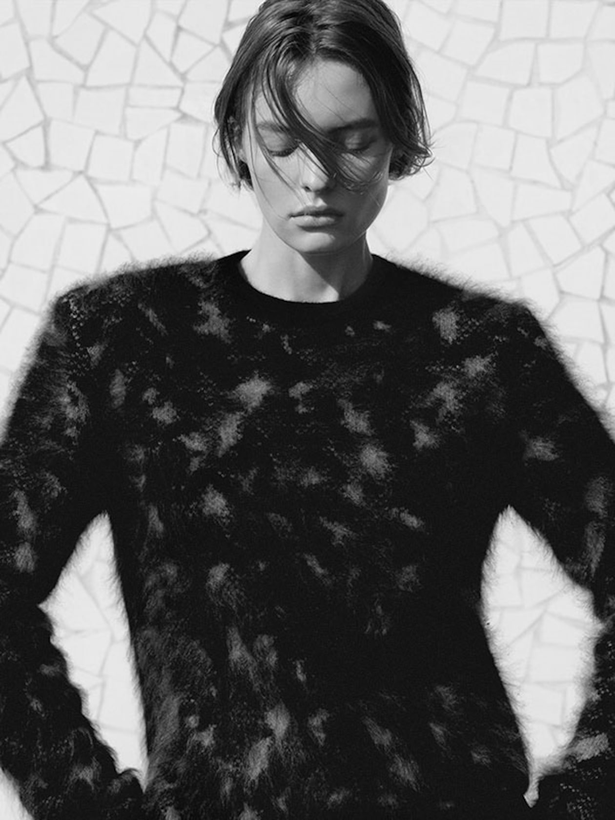 A person wearing a textured black sweater stands against a tiled wall, with eyes closed and hair gently blowing across the face.