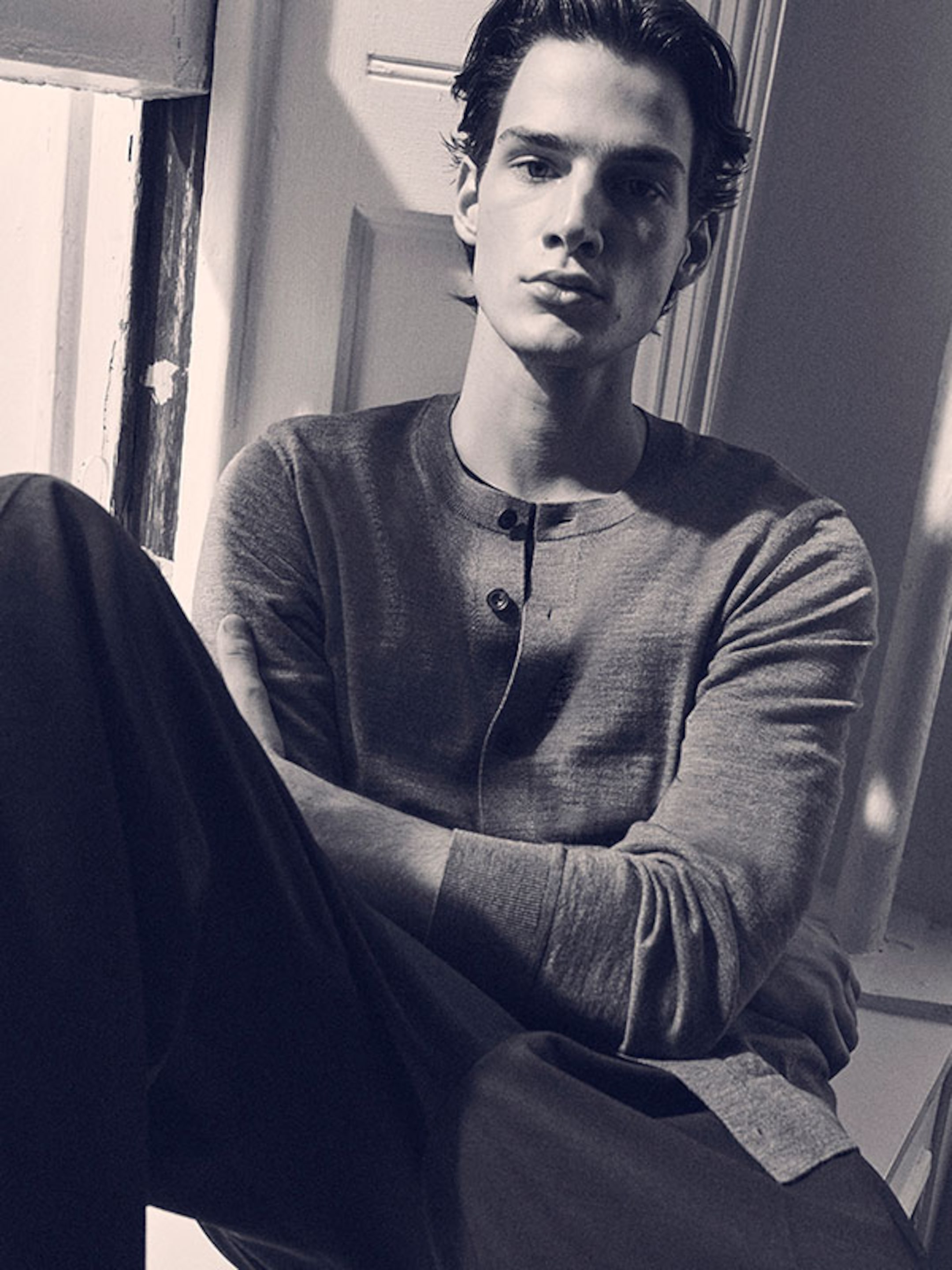 Man in a long-sleeve shirt sits by a window, arms crossed, looking at the camera in a monochrome setting.