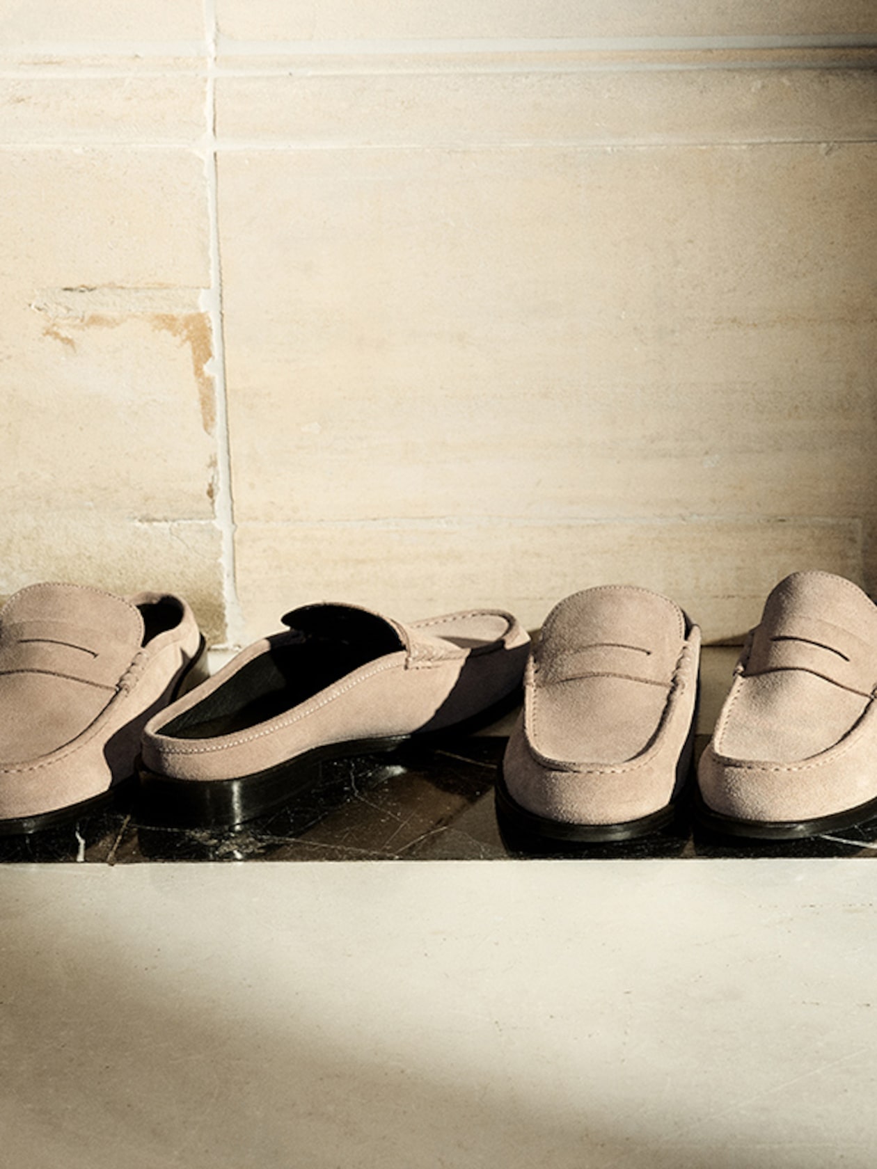 Two pairs of beige suede loafers on a marble floor, with sunlight casting shadows against a light-colored tiled wall.