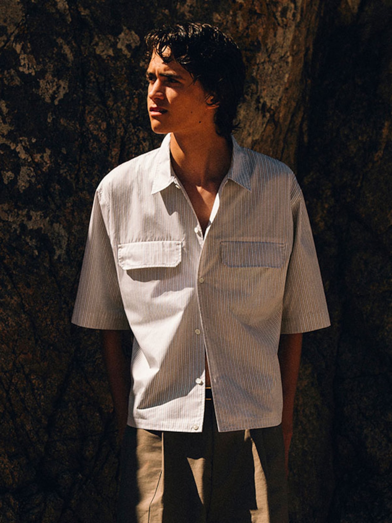 Young man in short-sleeve striped button-up shirt standing against a rocky backdrop, looking to the left in warm sunlight.