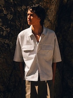 Placeholder for: Young man in short-sleeve striped button-up shirt standing against a rocky backdrop, looking to the left in warm sunlight.