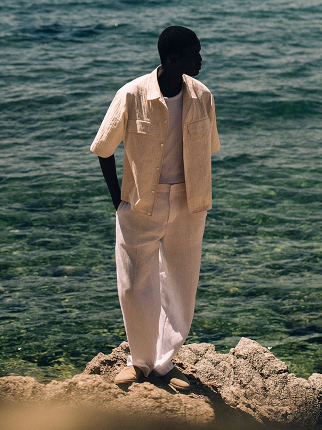 Silhouetted man in light linen outfit standing on rocky shore, looking toward the sea.