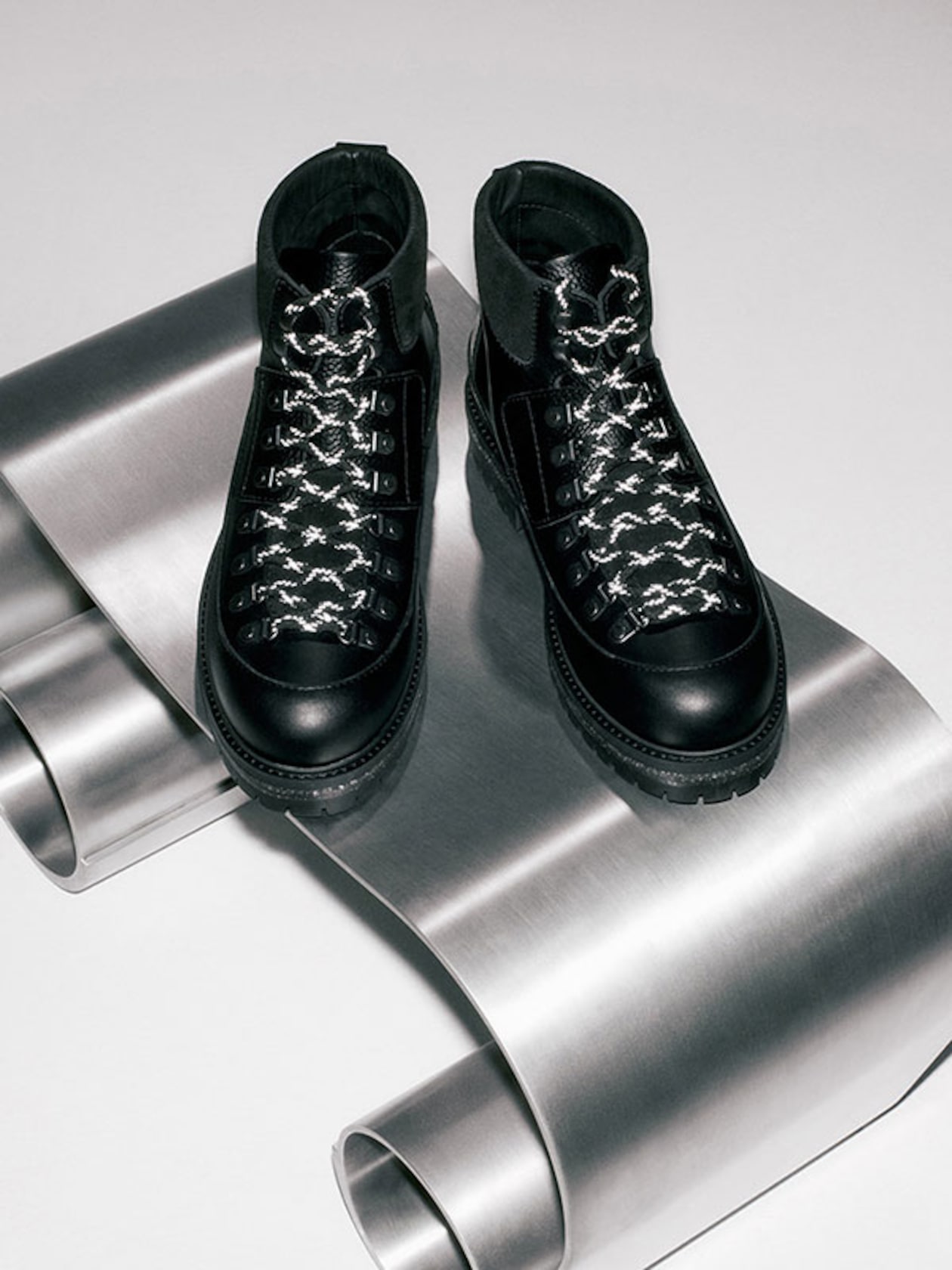 Black leather boots with patterned laces displayed on a curved metallic surface.