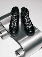 Placeholder for: Black leather boots with patterned laces displayed on a curved metallic surface.