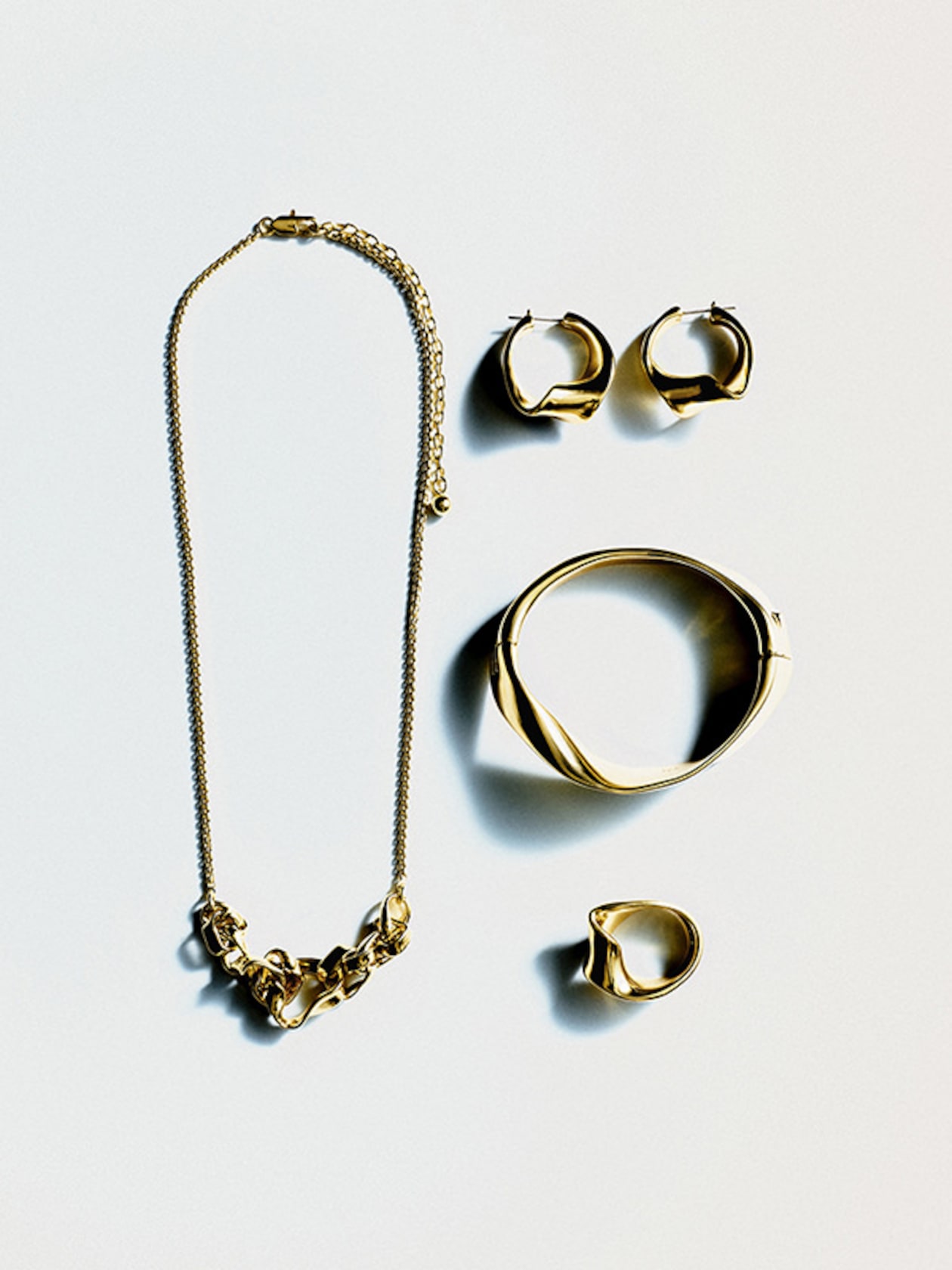 Gold jewelry set on a white background, featuring a chain necklace, hoop earrings, a bracelet, and a ring, all with a modern design.