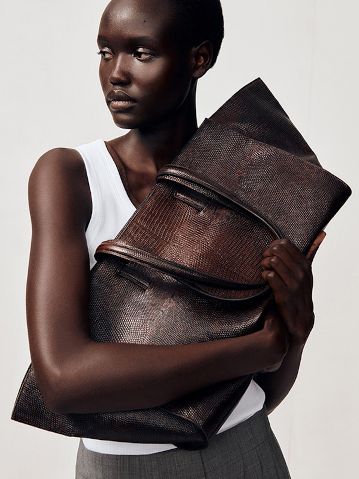 A person in a white tank top holds a large, textured brown leather bag, gazing to the side with a neutral expression.