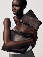 Placeholder for: A person in a white tank top holds a large, textured brown leather bag, gazing to the side with a neutral expression.