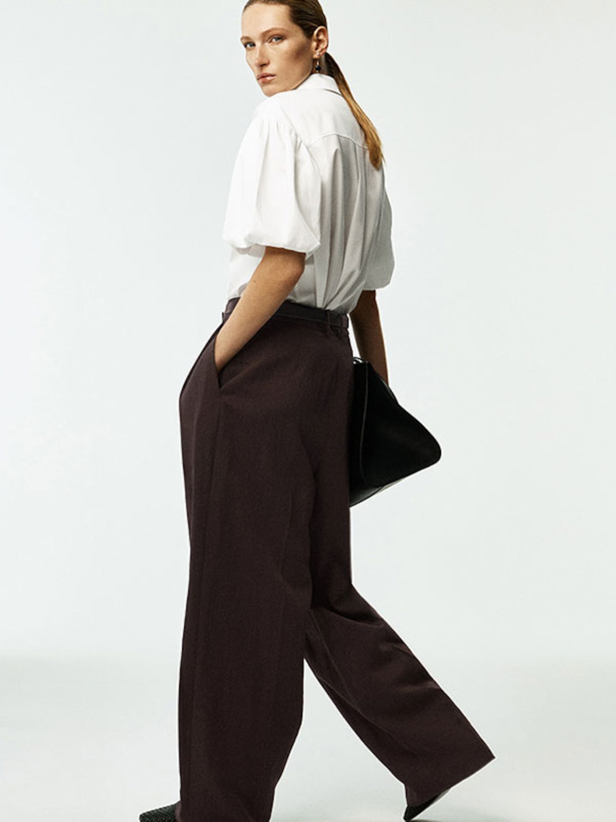 Person in a white blouse and brown wide-leg pants stands sideways, looking ahead, with hands in pockets, and a black bag.