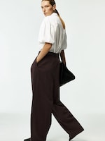 Placeholder for: Person in a white blouse and brown wide-leg pants stands sideways, looking ahead, with hands in pockets, and a black bag.