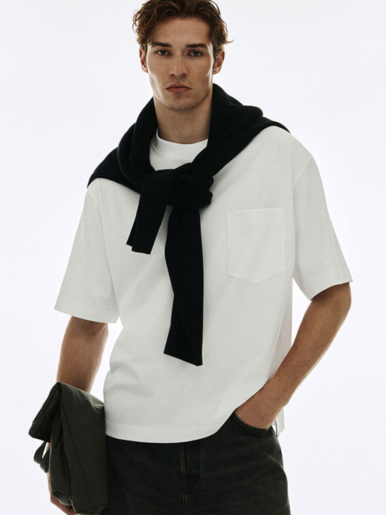 Man in a white t-shirt with a black sweater draped over shoulders, holding a dark jacket, against a light background.