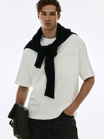 Placeholder for: Man in a white t-shirt with a black sweater draped over shoulders, holding a dark jacket, against a light background.