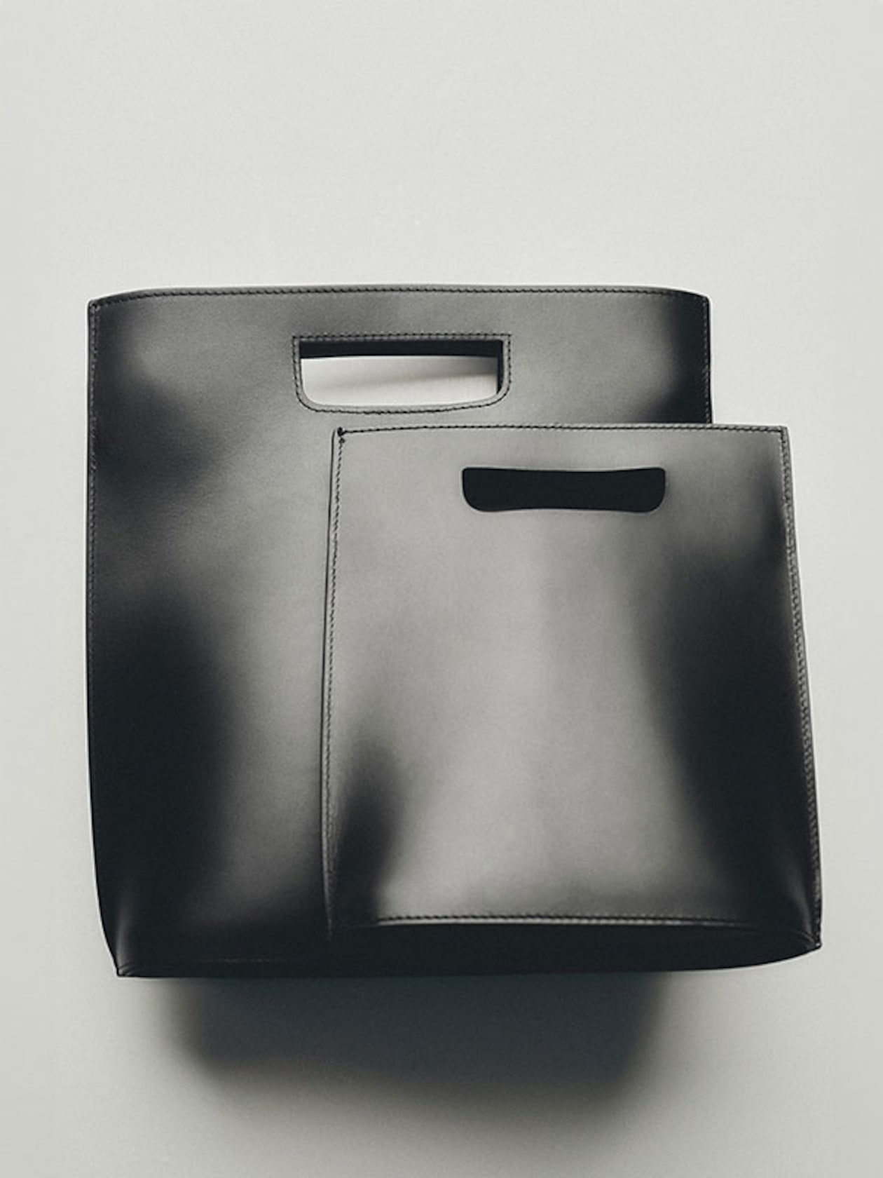 Two sleek black leather bags with handle cutouts, one slightly larger than the other, on a light gray background.