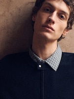 Placeholder for: A person with short hair wearing a dark jacket and a checkered shirt, posed against a textured wall, looking slightly upwards.