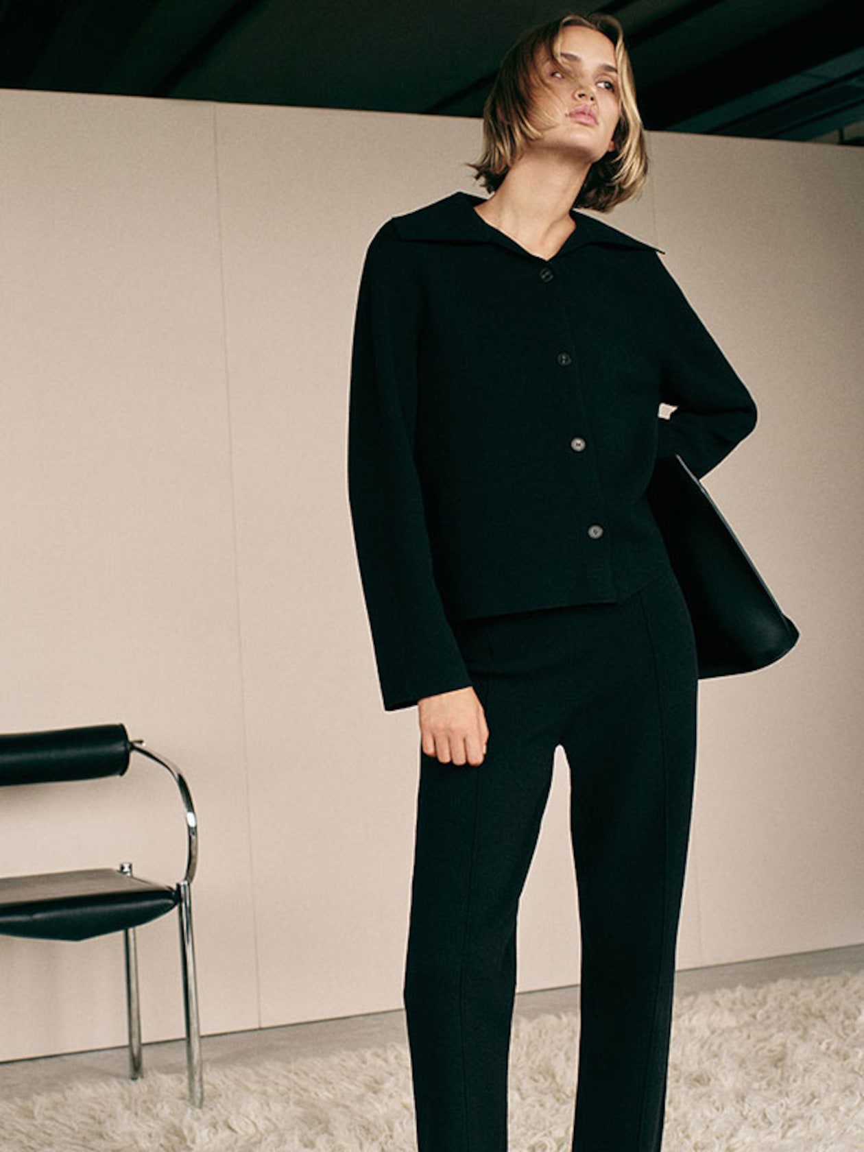 Model in a black tailored outfit stands confidently in a minimalist room with a leather chair and a shaggy white rug.