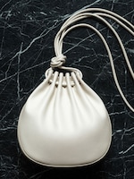 Placeholder for: Top-down view of a cream-colored, soft leather drawstring pouch bag resting on a dark green marbled surface. The bag features a gathered pleated top and a long, thin leather cord strap tied in a knot at the closure.