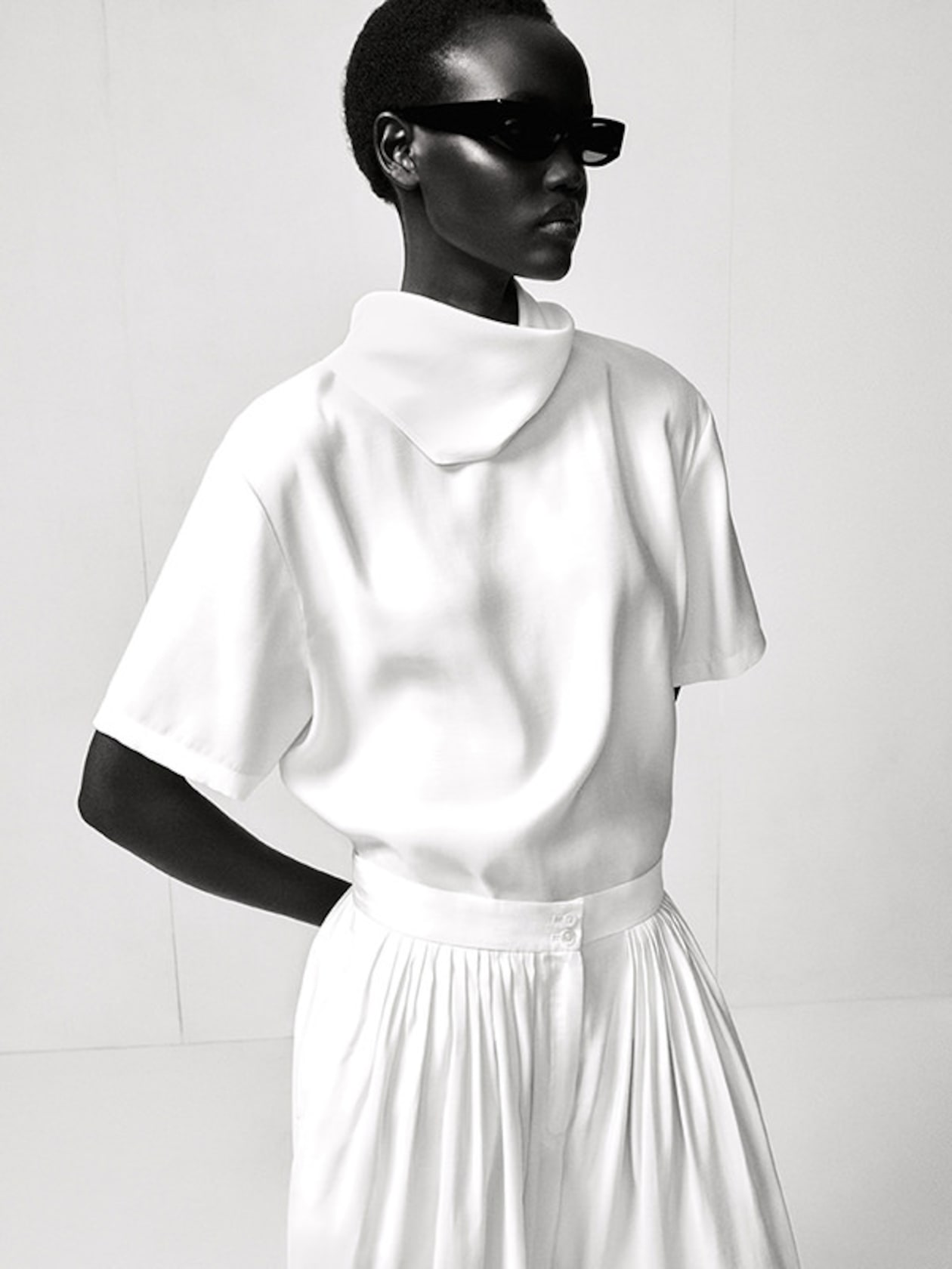 A model in a sleek white outfit with a high collar and pleated skirt, wearing dark sunglasses, poses against a plain backdrop.