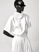Placeholder for: A model in a sleek white outfit with a high collar and pleated skirt, wearing dark sunglasses, poses against a plain backdrop.