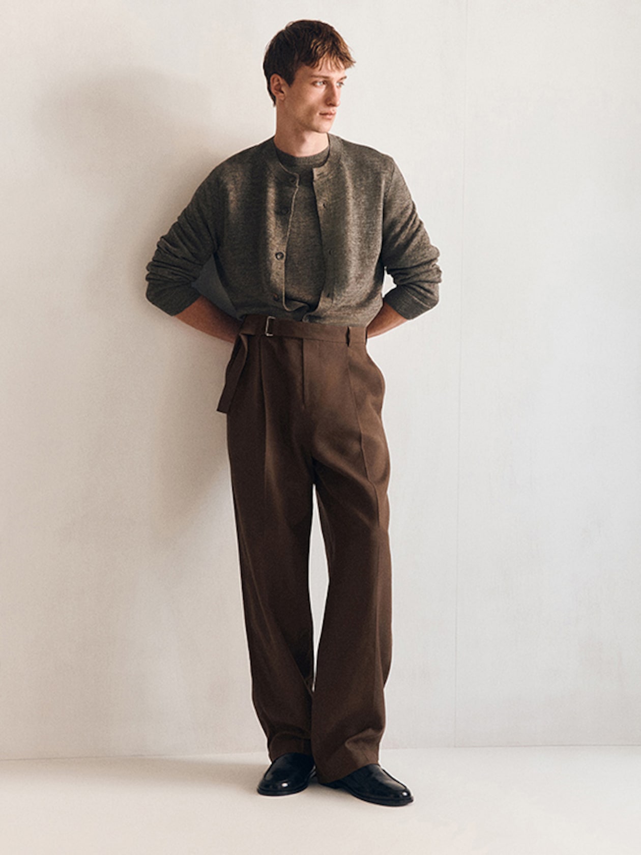 A person wearing a gray cardigan and loose brown trousers, standing casually against a plain white wall, with hands in pockets.