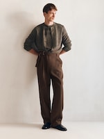 Placeholder for: A person wearing a gray cardigan and loose brown trousers, standing casually against a plain white wall, with hands in pockets.