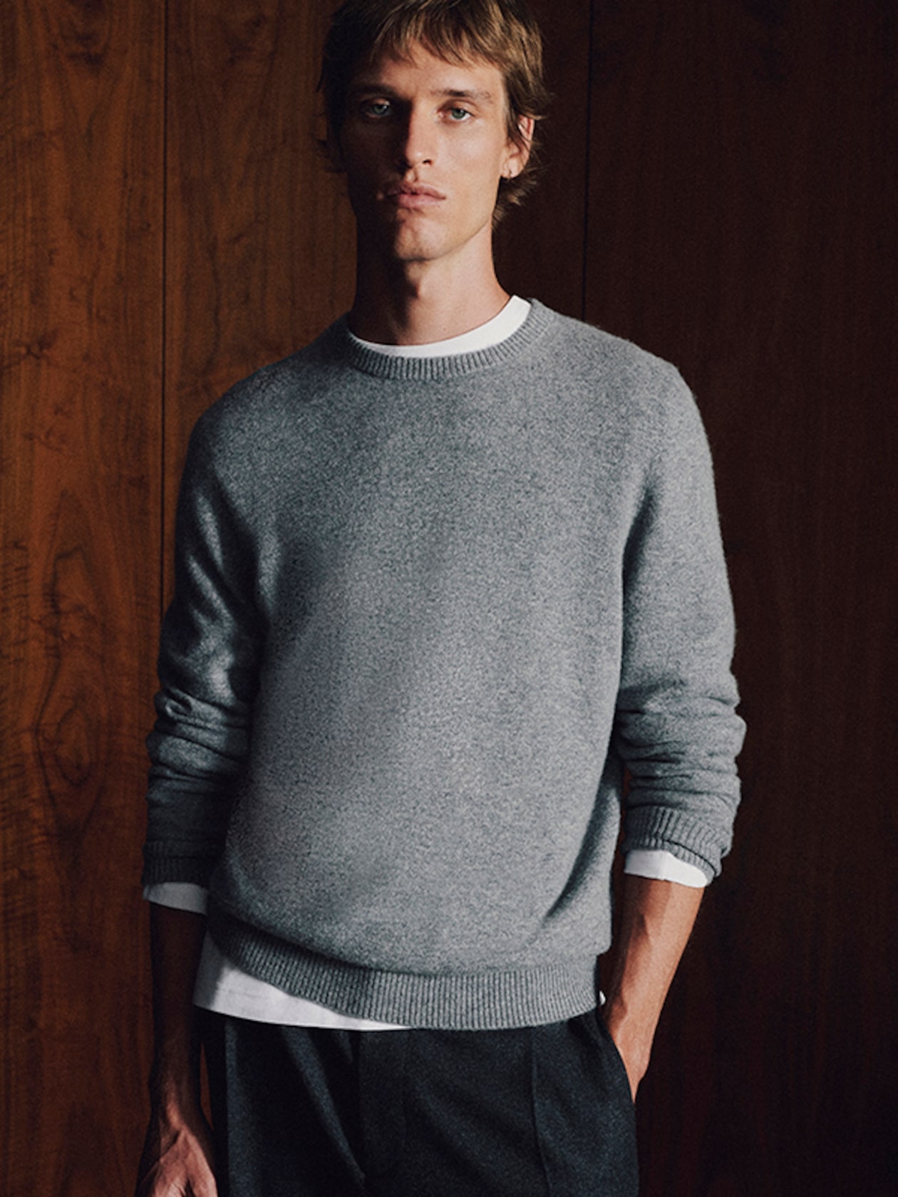 A person wearing a gray sweater and dark pants stands against a wooden wall, hands in pockets, looking forward.