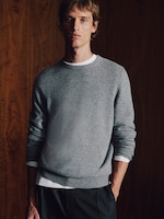 Placeholder for: A person wearing a gray sweater and dark pants stands against a wooden wall, hands in pockets, looking forward.
