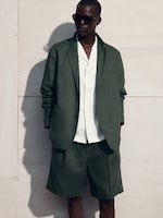 Placeholder for: Man in dark green suit with shorts and a white shirt, wearing sunglasses, stands against a light wall, casting a shadow.