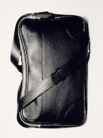Placeholder for: Black leather messenger bag with a textured surface and a long adjustable strap, placed against a light background.