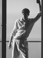 Placeholder for: A black-and-white lifestyle photograph of a man standing in a doorway overlooking the sea. He is wearing a relaxed-fit, long-sleeved white linen shirt with rolled-up sleeves and matching light-colored trousers with a thin belt. The lighting is soft and atmospheric, highlighting the natural texture of the linen fabric.