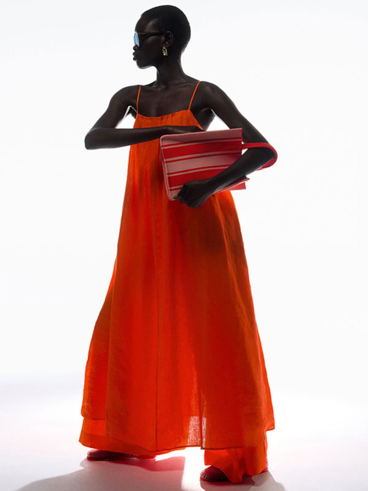 Person in an orange dress holds a red and white striped clutch, wearing sunglasses and earrings, standing against a white background.
