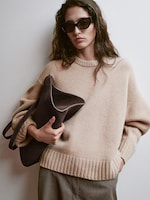 Placeholder for: Woman in a beige sweater, wearing large sunglasses, holds a dark suede bag. Her hair is loose, and she stands against a neutral background.