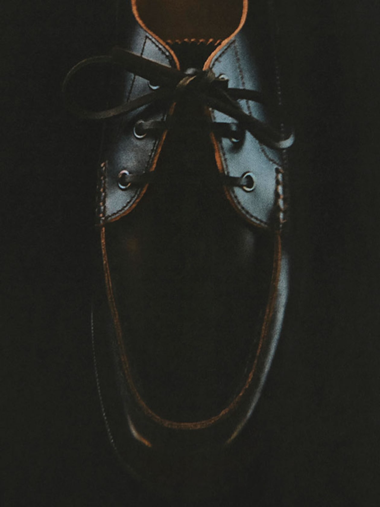 A dark leather shoe with laces, partially lit, highlighting its smooth texture and stitching details against a black background.