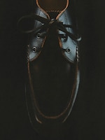 Placeholder for: A dark leather shoe with laces, partially lit, highlighting its smooth texture and stitching details against a black background.