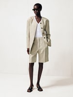 Placeholder for: Person wearing a beige oversized blazer, matching bermuda shorts, white top, black sandals, and sunglasses, standing against a light background.