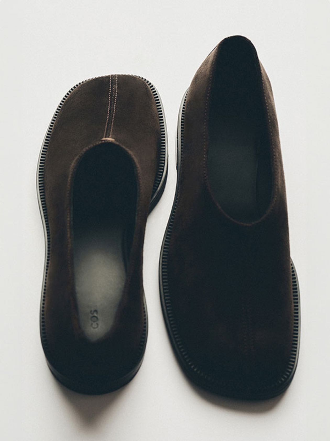 Pair of dark brown suede slip-on shoes on a light background, showing top and bottom views with visible stitching and textured soles.