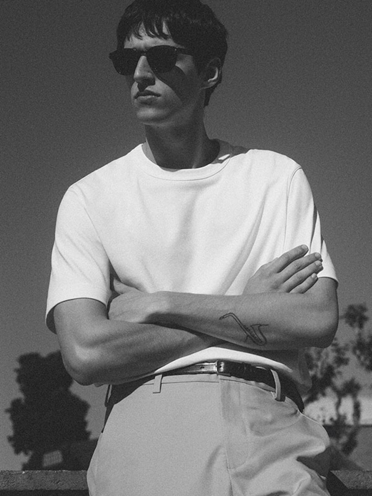 A black-and-white portrait of a man with arms crossed, wearing a clean-cut white short-sleeved T-shirt tucked into tailored trousers. The outfit is accessorized with a slim dark leather belt and classic dark sunglasses. A small saxophone tattoo is visible on his forearm against a clear sky background.