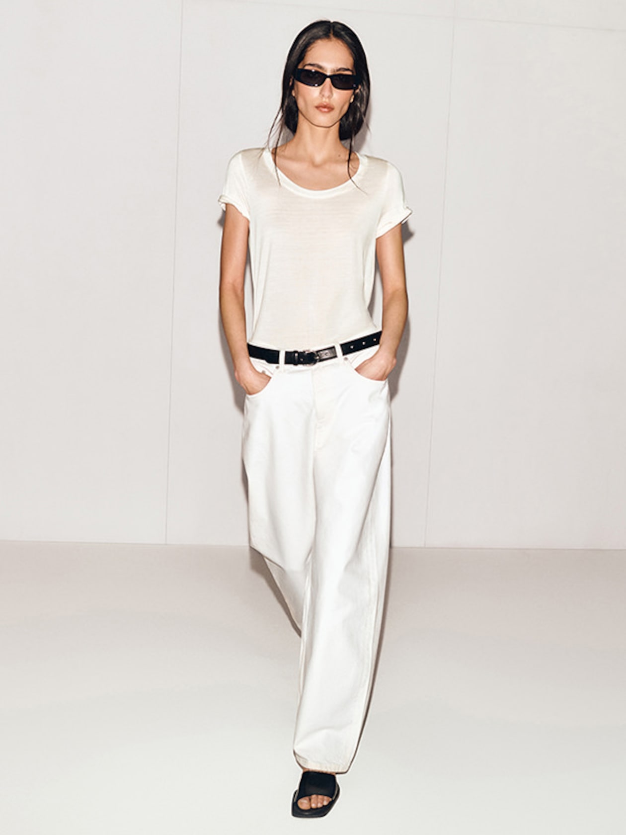 A woman in sunglasses wearing a white T-shirt, loose white pants, and black sandals, stands with hands in pockets against a plain background.