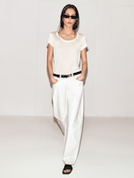 Placeholder for: A woman in sunglasses wearing a white T-shirt, loose white pants, and black sandals, stands with hands in pockets against a plain background.