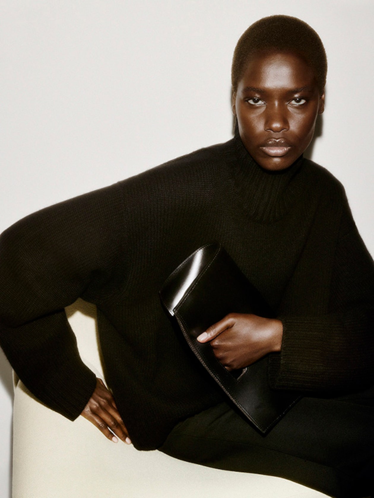 Person in a black sweater seated against a white background, holding a black clutch, with a confident expression.