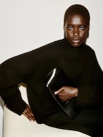 Placeholder for: Person in a black sweater seated against a white background, holding a black clutch, with a confident expression.