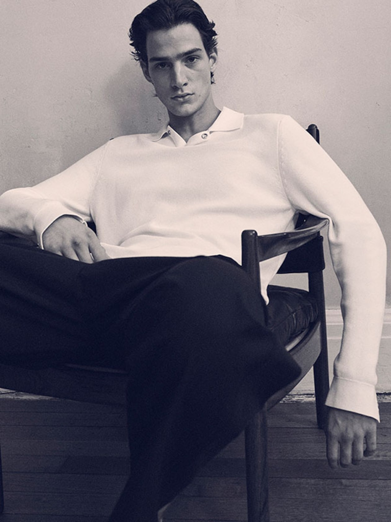 Male model sitting in a chair wearing a white sweater and dark pants, with a relaxed posture and neutral expression.