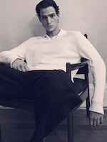 Placeholder for: Male model sitting in a chair wearing a white sweater and dark pants, with a relaxed posture and neutral expression.