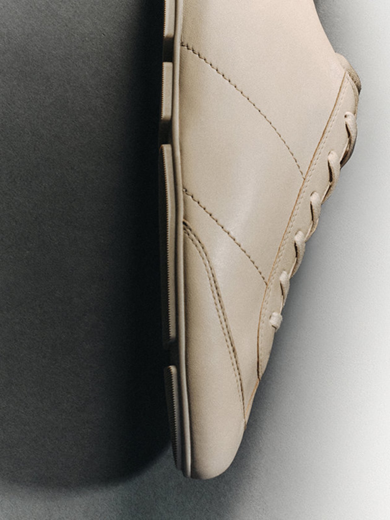Beige leather sneaker viewed from the side on a two-toned background, showing details of stitching and laces.