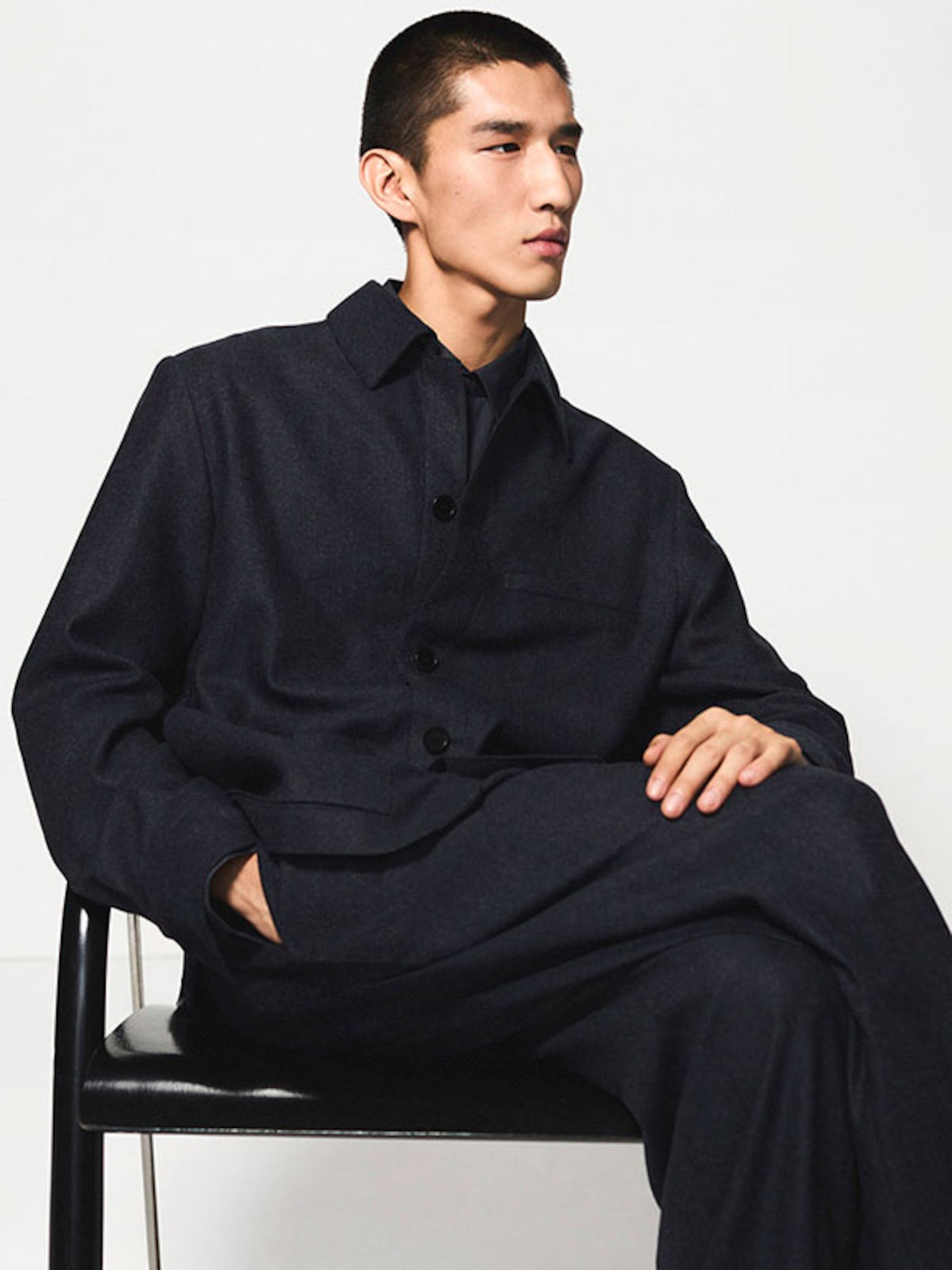 Person in a dark button-up jacket and matching pants, sitting casually on a black chair against a white background.