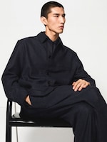 Placeholder for: Person in a dark button-up jacket and matching pants, sitting casually on a black chair against a white background.