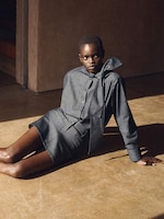 Placeholder for: Model in a dark grey outfit sits on the floor, leaning back with one arm supporting their weight, in a sunlit room.