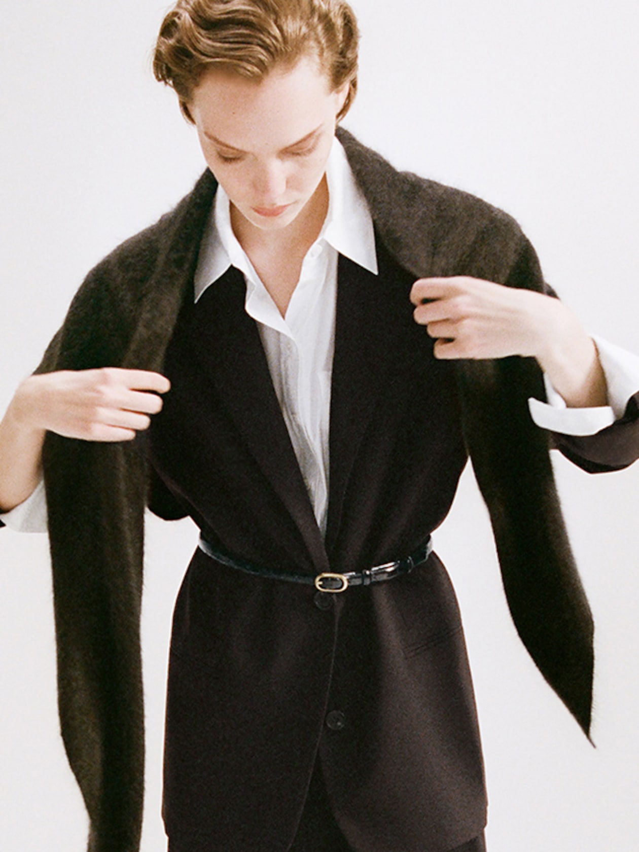 Person in a black suit and white shirt, adjusting a brown scarf around their shoulders, with a black belt accentuating the waist.